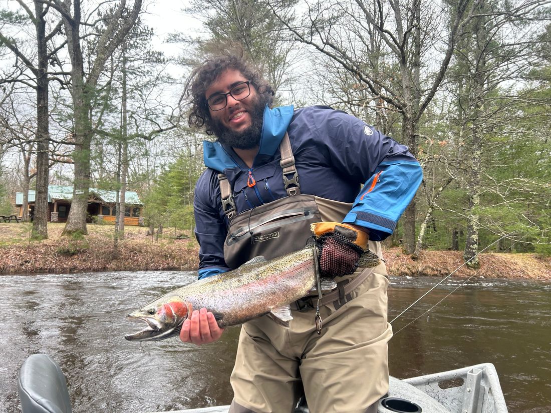 Gabriel holding a steelhead in a drift boat on a forested river