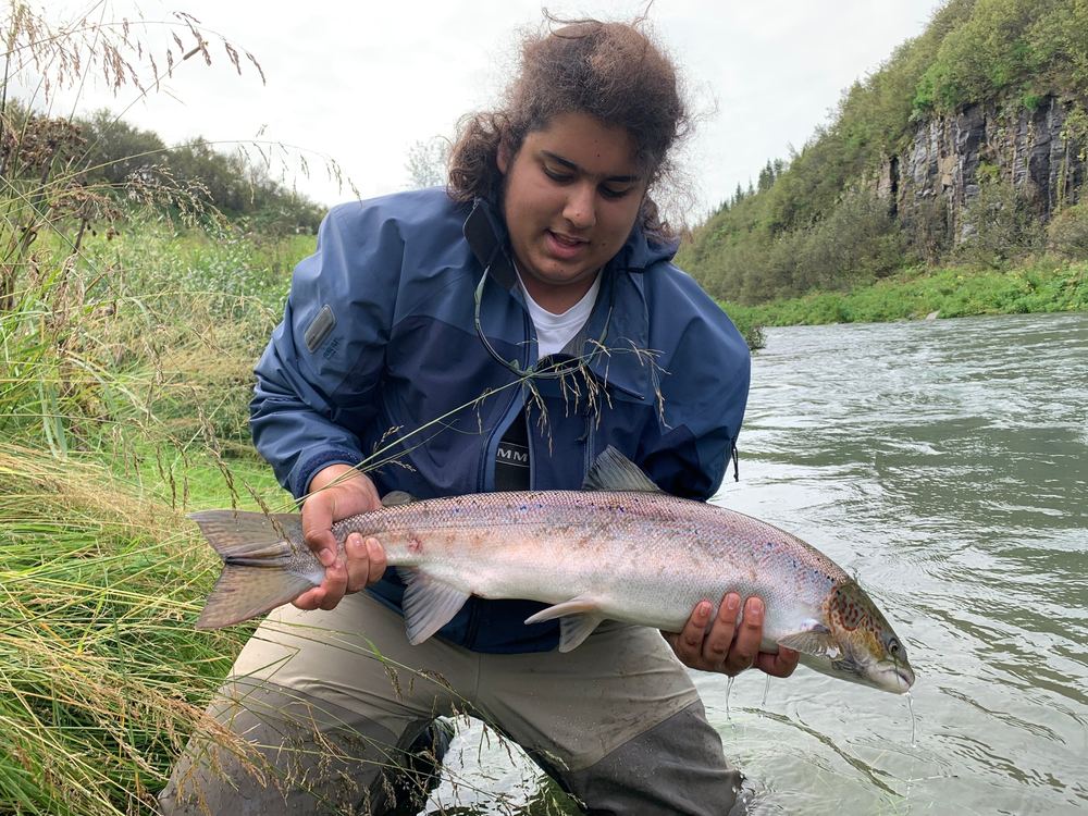 Gabriel kneeling in a river holding a large Atlantic salmon, grassy bank and a cliff behind