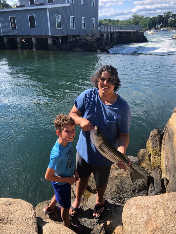 Gabriel holding a striped bass on harbor rocks beside a boy, a building on pilings and rushing water behind