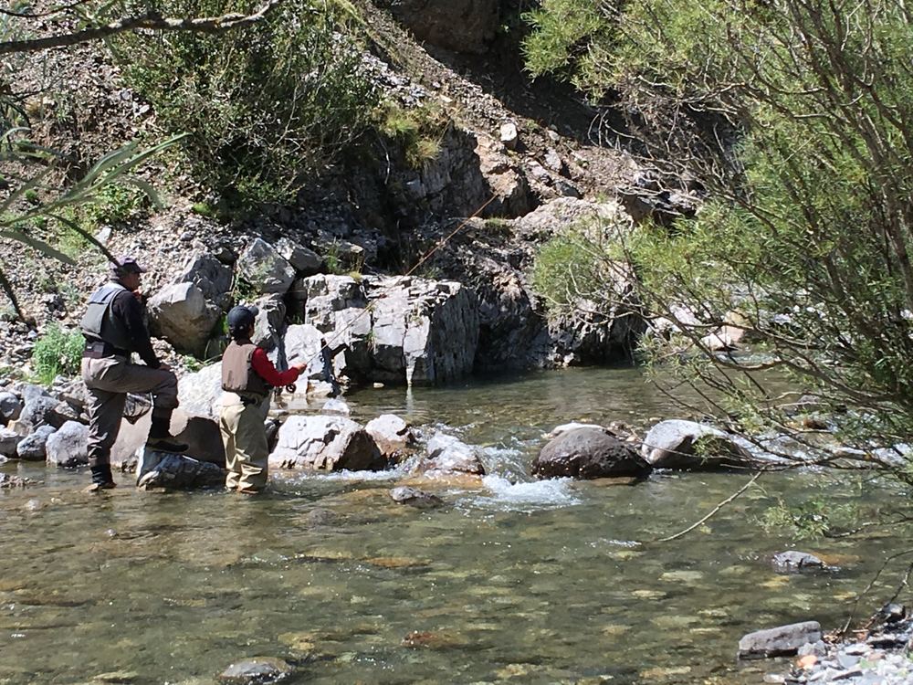 Two anglers in waders on a clear, rocky mountain stream, one casting a fly rod toward a rock wall