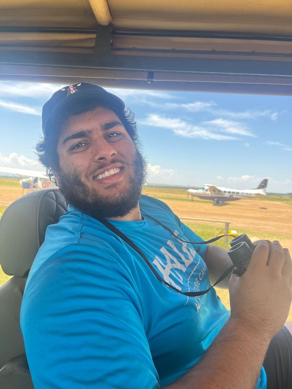 Gabriel in a safari vehicle holding binoculars, small planes on an airstrip behind him